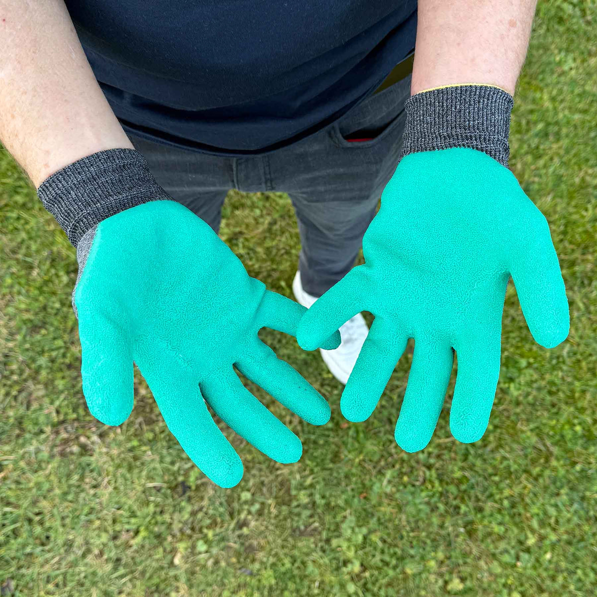 Person zeigt beide Hände mit grünen, latexbeschichteten Gartenhandschuhen über einer Rasenfläche.