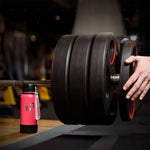 Pinke Trinkflasche mit Trageschlaufe trägt einer schwer beladenen Langhantel im Fitnessstudio, während eine Hand eine zusätzliche Gewichtsscheibe auf die Stange schiebt.