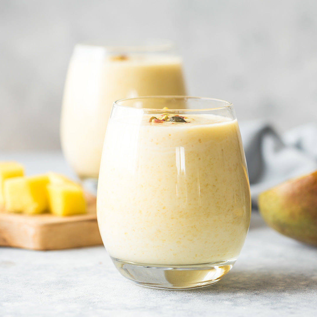 Zwei Gläser mit cremigem, hellgelbem Fruchtshake, davor Mangostücke auf einem Holzbrett.