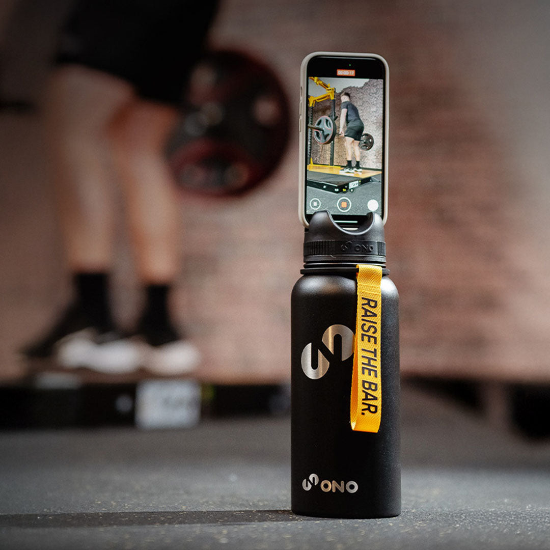 Schwarze Trinkflasche mit gelber Schlaufe und Deckel als Smartphone-Halter steht im Fitnessstudio; auf dem Smartphone wird eine Gewichtheber-Übung gefilmt, im Hintergrund ist unscharf eine Person mit Langhantel zu sehen.