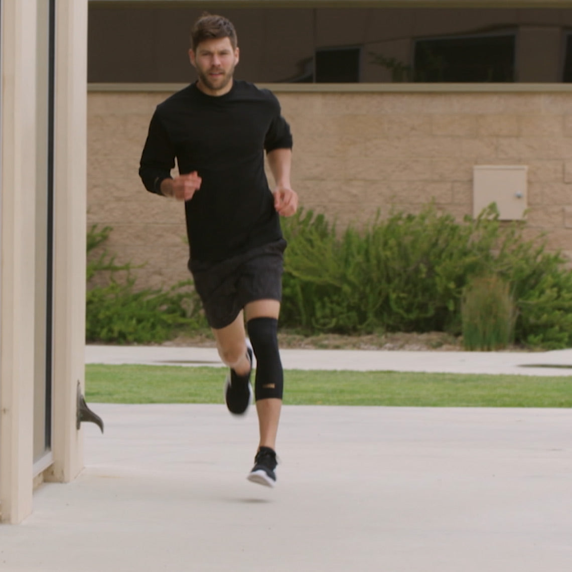 in Mann joggt im Freien auf einem gepflasterten Weg. Er trägt Sportkleidung in Schwarz und eine schwarze Kniebandage am rechten Bein. Im Hintergrund sind Büsche und eine helle Gebäudewand zu sehen.