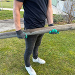 Person mit grün-grauen Gartenhandschuhen hebt einen dicken Holzstamm auf einer Rasenfläche an.