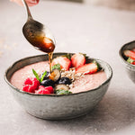 Smoothie-Bowl mit Erdbeeren, Himbeeren, Blaubeeren und Haferflocken, über die Honig von einem Löffel geträufelt wird.