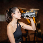 Sportliche Frau trinkt im Fitnessstudio aus einer schwarzen Trinkflasche mit Logo und gelber Schlaufe, während Trainingsgeräte unscharf im Hintergrund zu sehen sind.