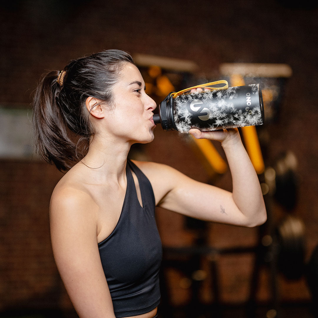Sportliche Frau trinkt im Fitnessstudio aus einer schwarzen Trinkflasche mit Logo und gelber Schlaufe, während Trainingsgeräte unscharf im Hintergrund zu sehen sind.