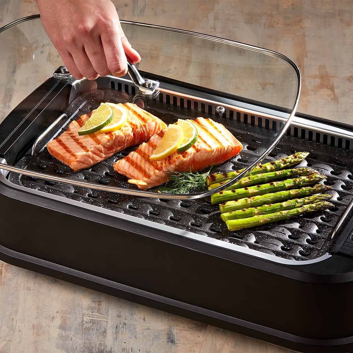 PowerXL Smokeless Grill – gegrillter Lachs mit Limette und Spargel unter Glasdeckel auf gerillter Platte.