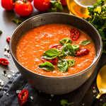 Schüssel mit frischer Gazpacho, garniert mit Basilikumblättern und roten sowie grünen Chilischeiben, umgeben von Tomaten und Gewürzen.
