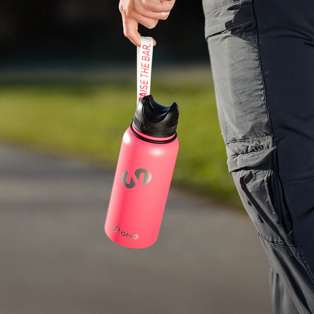 Pinke Trinkflasche mit schwarzem Deckel und Trageschlaufe wird an der Schlaufe neben dem Bein einer Person im Freien gehalten.