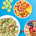 Verschiedene frisch zubereitete Gerichte, darunter Gurkensalat, bunte Obst-Bowl und würzige Mango-Salsa, angerichtet auf blauem Hintergrund