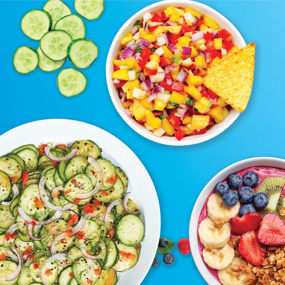 Verschiedene frisch zubereitete Gerichte, darunter Gurkensalat, bunte Obst-Bowl und würzige Mango-Salsa, angerichtet auf blauem Hintergrund