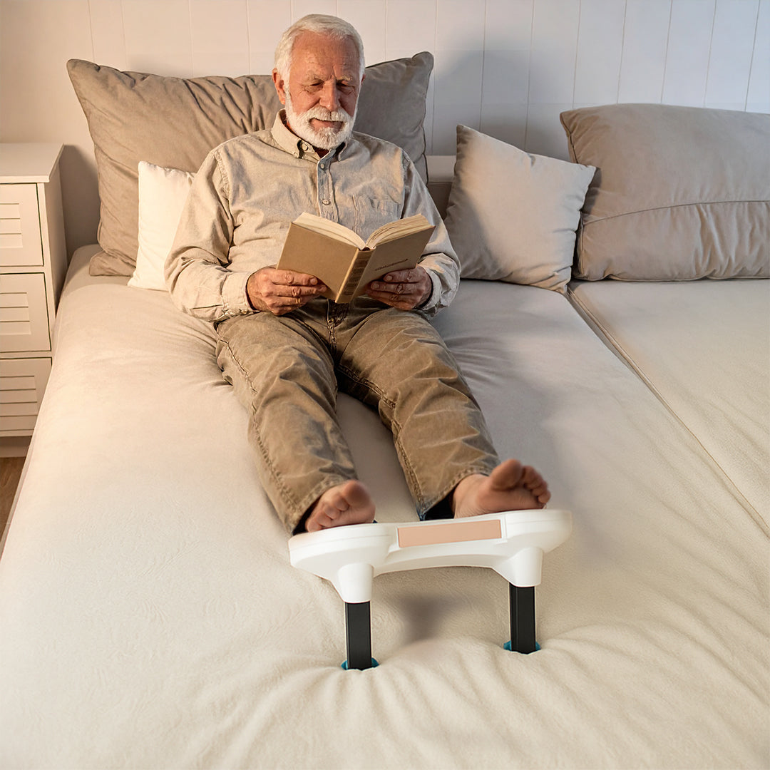 Älterer Mann liegt im Bett, liest ein Buch und nutzt dabei das Fllow Fußmassagegerät zur Entspannung.