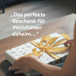 Hände halten eine dekorative Geschenkbox mit goldener Schleife und dem Text ‚Das perfekte Geschenk für Wohlfühlen daheim