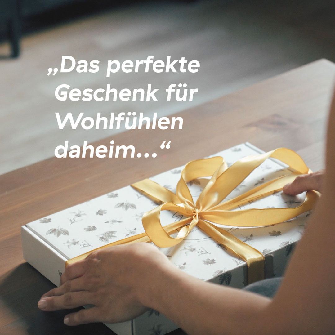 Hände halten eine dekorative Geschenkbox mit goldener Schleife und dem Text ‚Das perfekte Geschenk für Wohlfühlen daheim