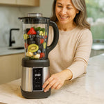 Lächelnde Frau in einer Küche bedient einen Standmixer, der mit frischen Früchten wie Beeren, Kiwi und Bananen gefüllt ist.