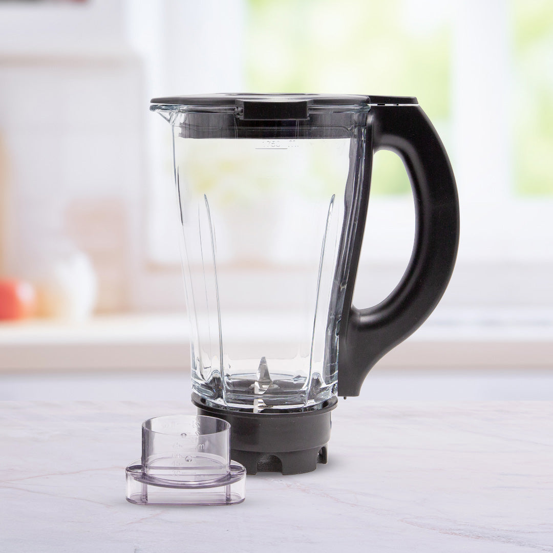 Leerer transparenter Glasbehälter-Mixaufsatz mit schwarzem Griff steht auf einer Küchenarbeitsplatte, daneben der abgenommene Messbecherdeckel.