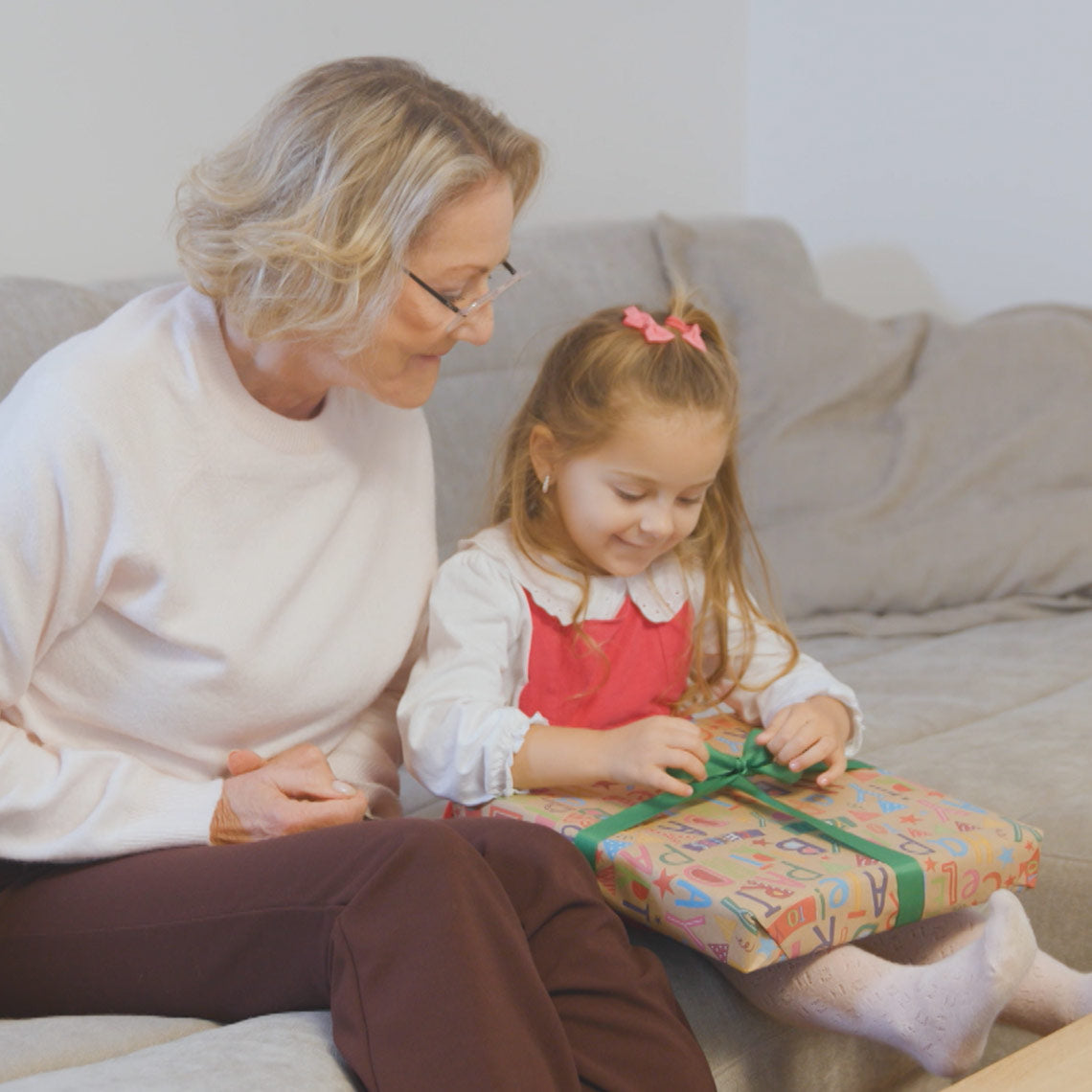 Mädchen öffnet ein Geschenk neben einer älteren Frau auf dem Sofa.