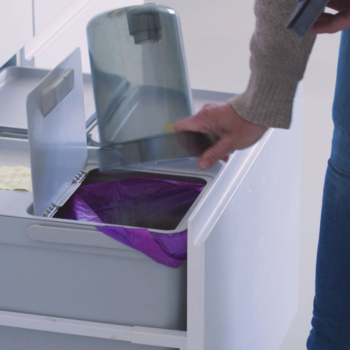 Eine Hand leert den transparenten Staub- und Schmutzbehälter eines Geräts in einen geöffneten Mülleimer mit Müllbeutel. Der Behälter wird zum Entleeren über den Abfall gehalten, um den gesammelten Schmutz hygienisch zu entsorgen.