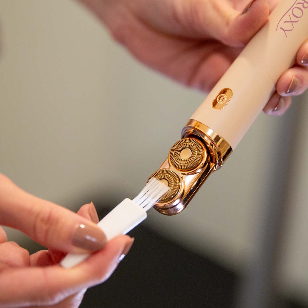 Nahaufnahme eines elektrischen Beauty-Geräts mit goldfarbenem Aufsatz, dessen Scherkopf mit einer kleinen Reinigungsbürste sorgfältig gereinigt wird.
