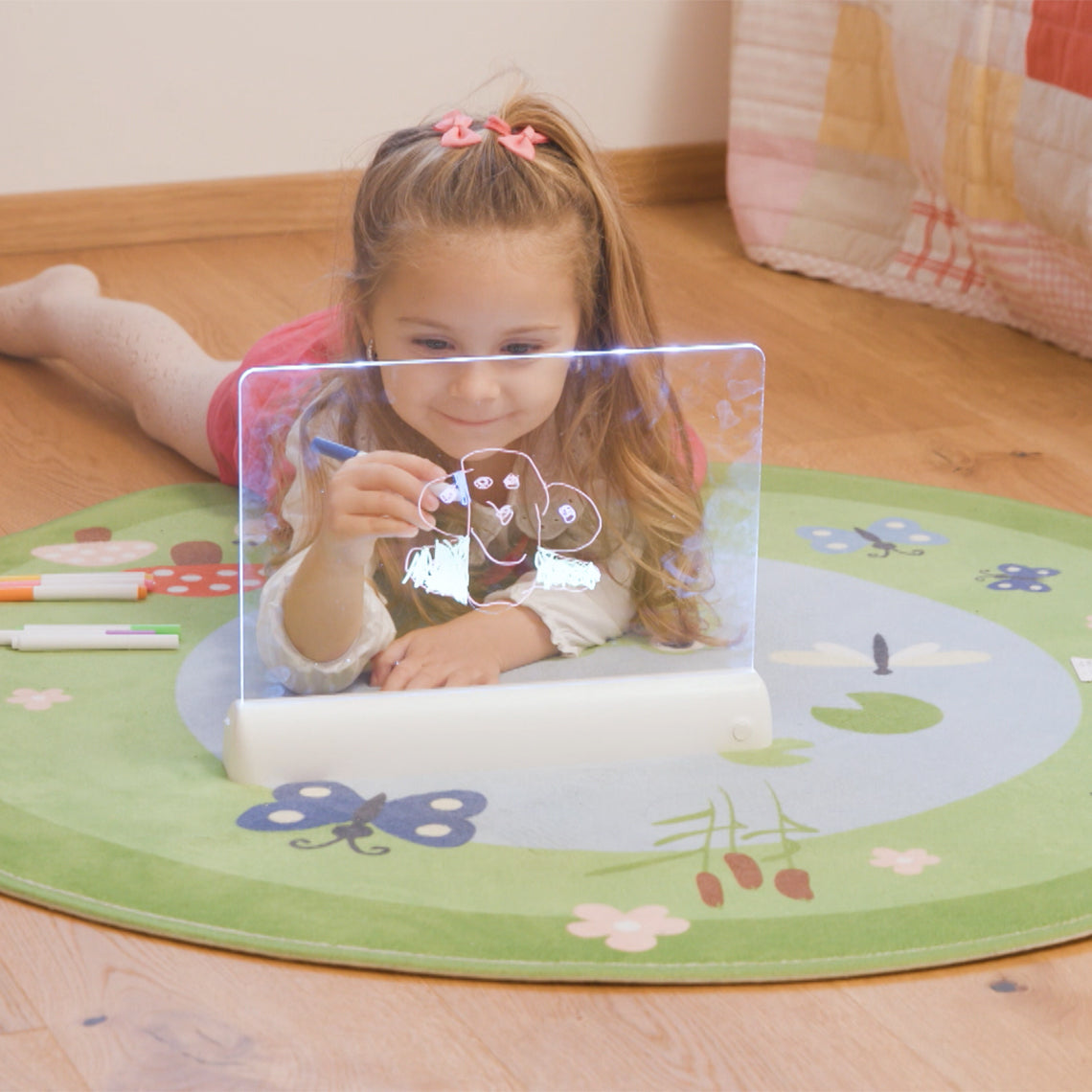 Mädchen liegt auf einer Spielmatte und zeichnet auf einer beleuchteten Mini-Leuchttafel.