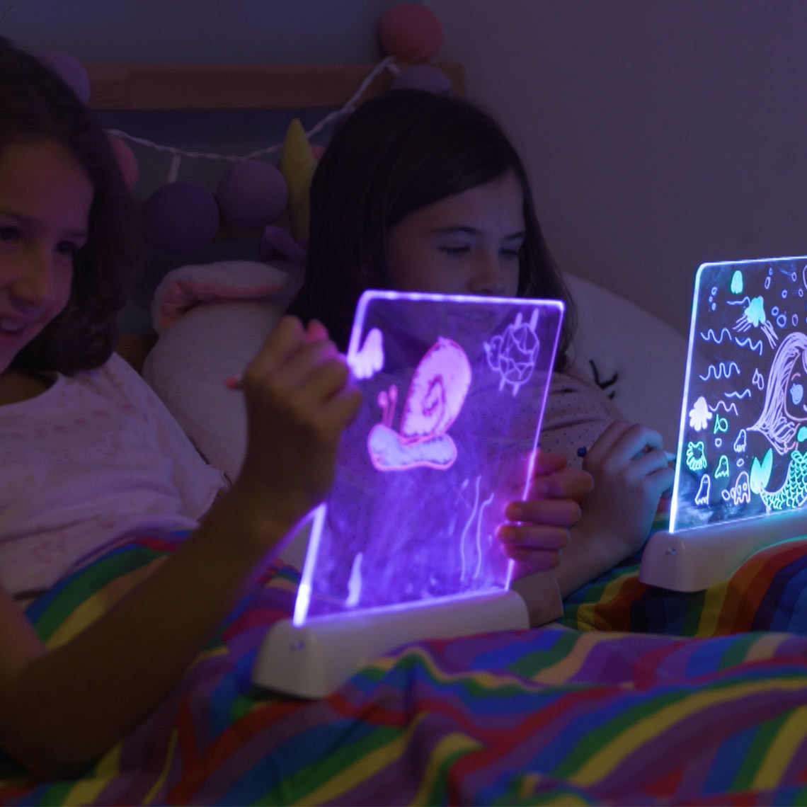 Zwei Mädchen zeichnen im Bett auf beleuchteten Mini-Leuchtboards mit buntem Lichteffekt.