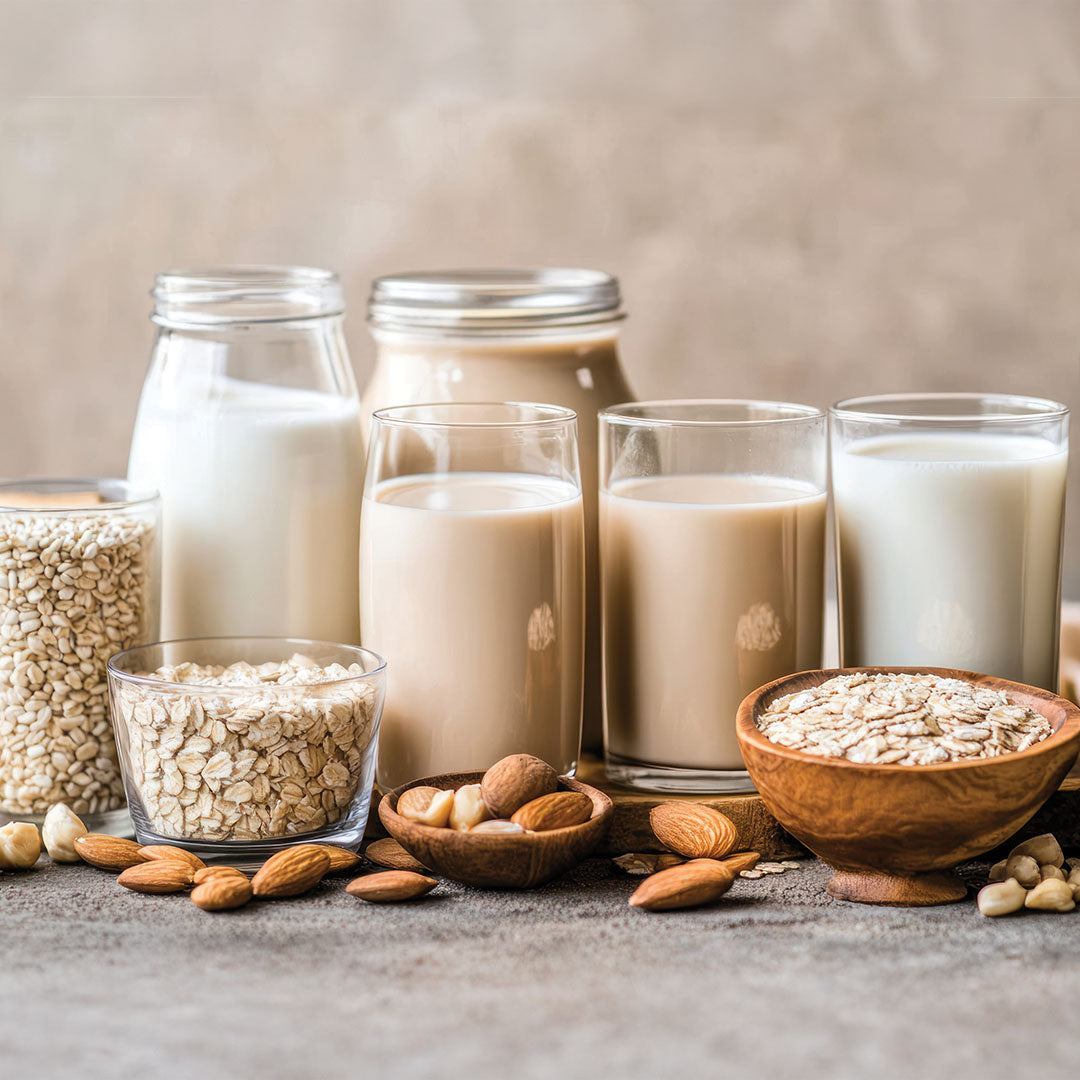 Verschiedene pflanzliche Milchsorten und Zutaten wie Hafer und Mandeln in Gläsern arrangiert.