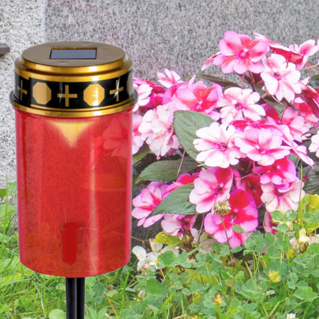 Rote Grabkerze mit schwarzem Golddeckel steht im Außenbereich neben blühenden Blumen.