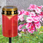Rote Grabkerze mit schwarzem Golddeckel steht im Außenbereich neben blühenden Blumen.