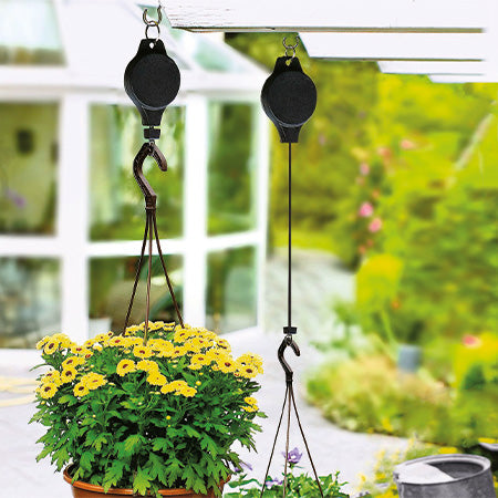 Zwei schwarze Pflanzenlifte mit Haken, an einer Pergola montiert, heben einen Blumentopf mit gelben Blumen.