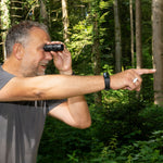 Mann zeigt lächelnd in den Wald, während er durch ein Taschenfernglas blickt.