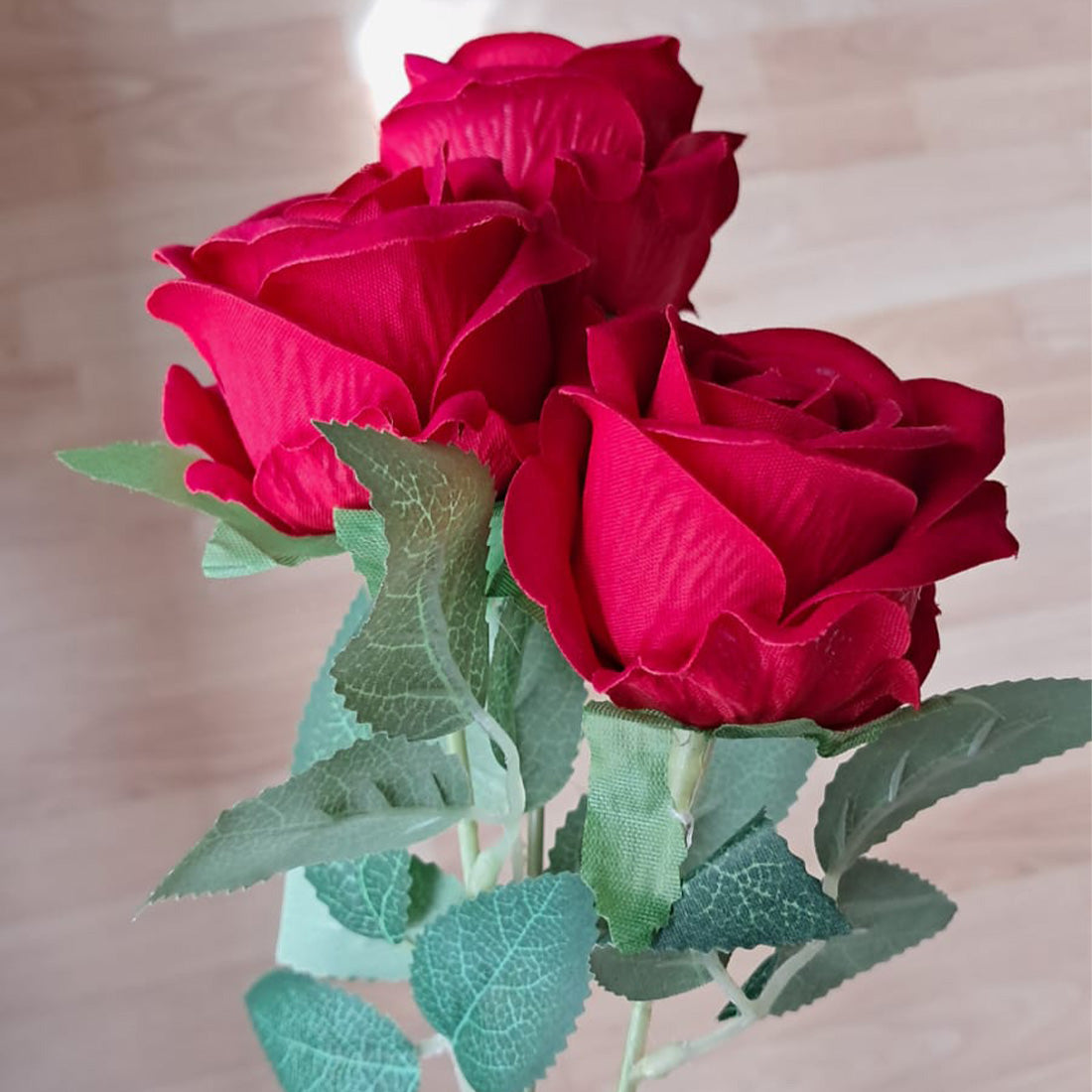 Drei künstliche rote Rosen aus Stoff mit grünen Blättern auf einer Holzoberfläche.