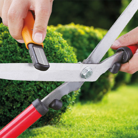 Ein 6in1-Messerschärfer schärft eine Gartenschere vor grünem Heckenhintergrund.